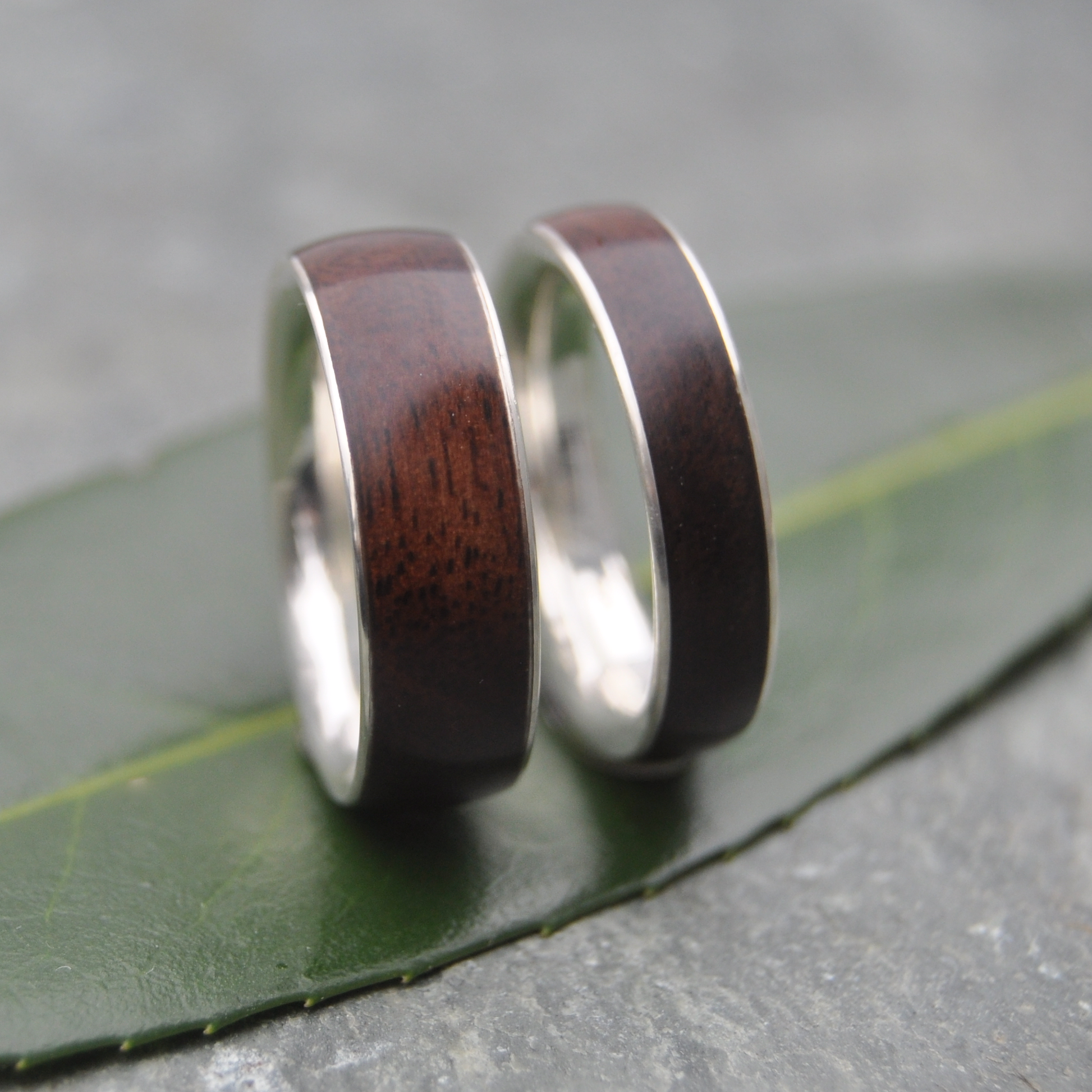 Two rings with dark wood and silver metal are placed on a green leaf.