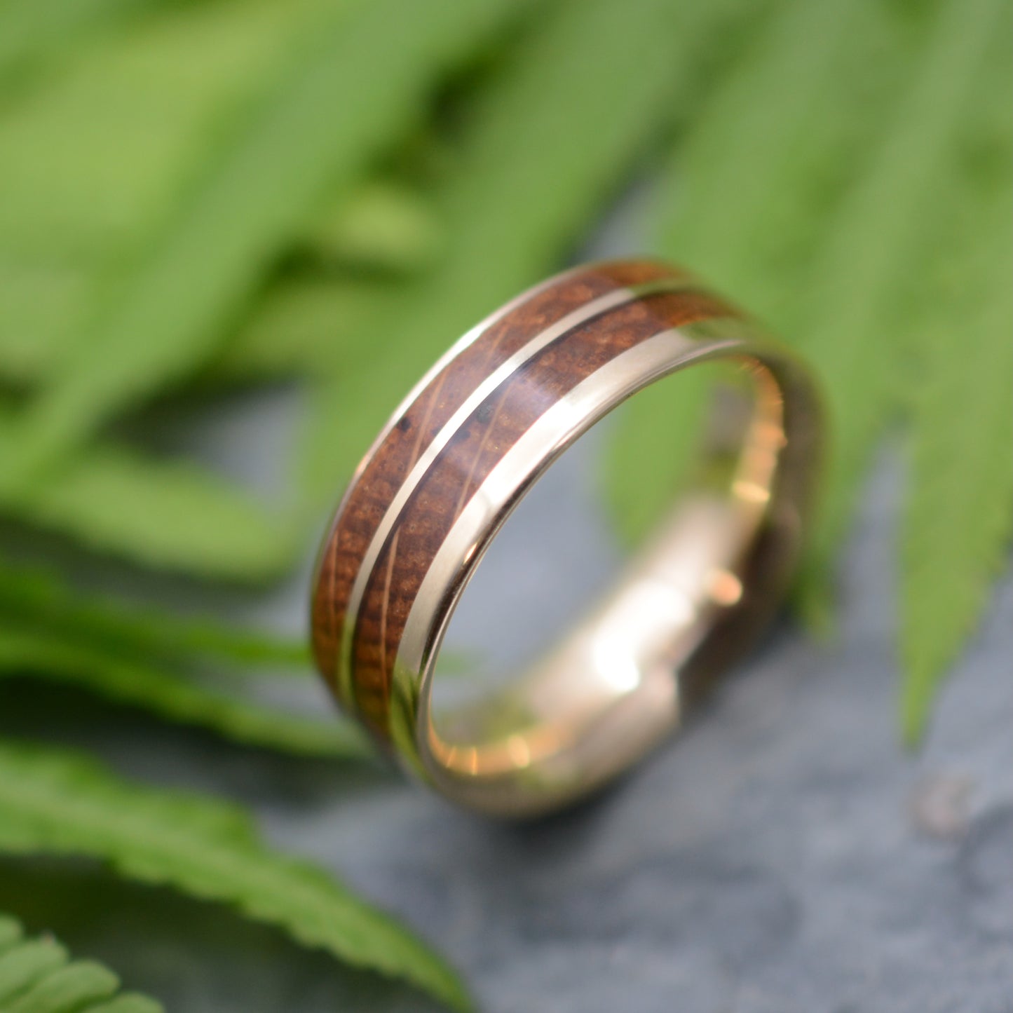 A gold ring with a wooden band is displayed against a blurred green background, with a leaf visible in the foreground.