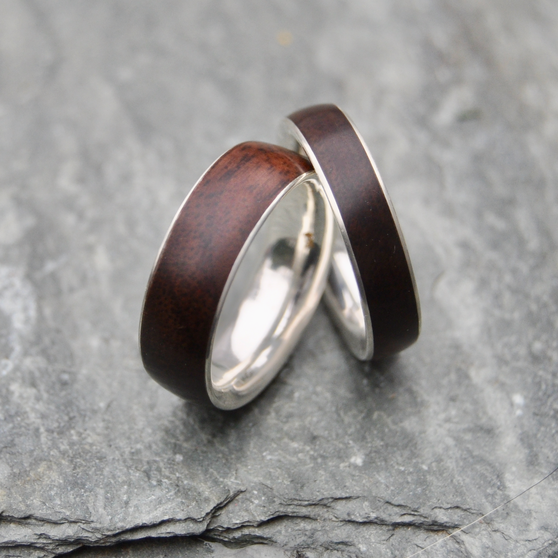 Two rings with dark wood and silver bands are placed on a gray rock surface.