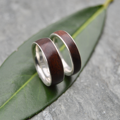 Two rings with dark wood and silver bands are placed on a green leaf against a gray background.