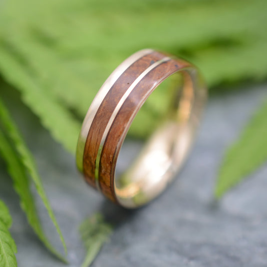 A close-up image of a wooden ring with a gold band, resting on a surface with green leaves in the background.