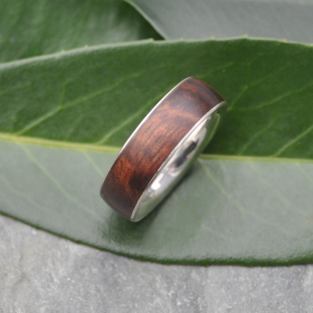 A wooden ring with a silver band is resting on a green leaf.